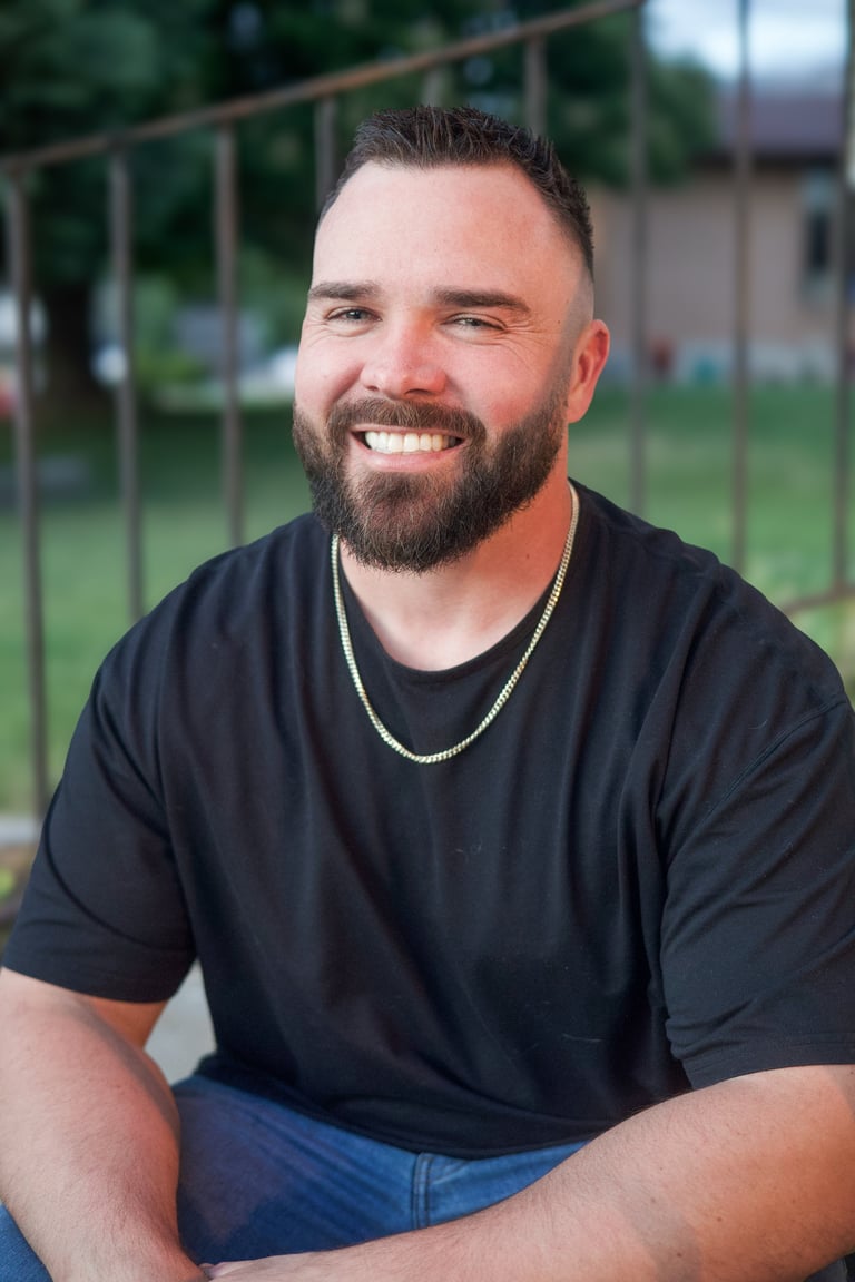 Smiling man with beard wearing black t-shirt and chain necklace, seated outdoors with fence in background