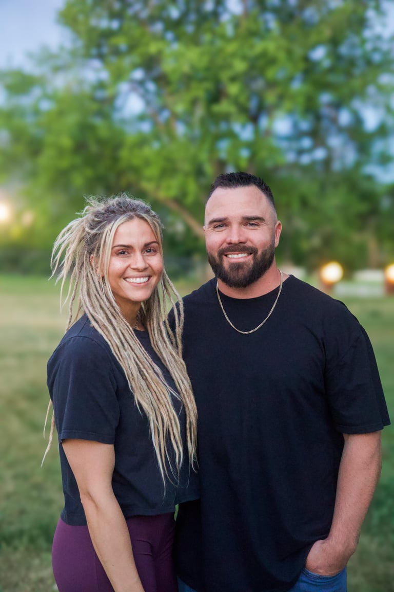 A smiling couple standing together outdoors in front of trees at dusk, both wearing black shirts