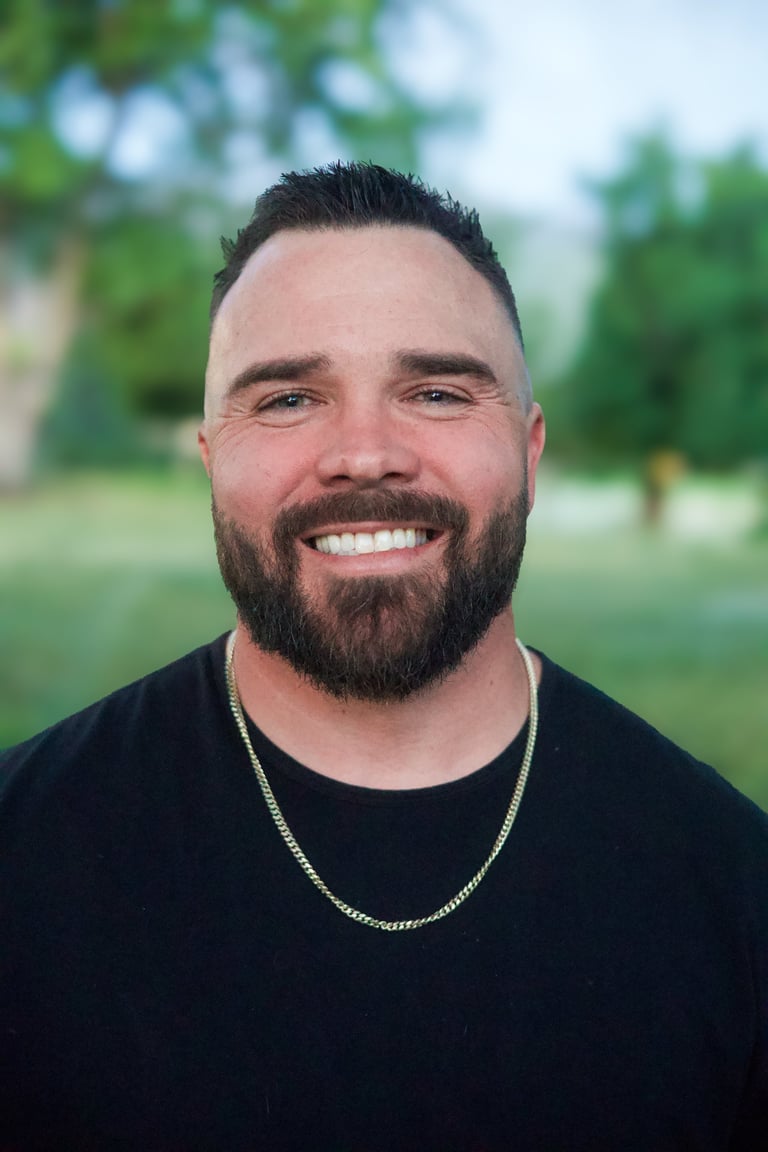 Portrait of a smiling man with a beard wearing a black shirt and chain necklace outdoors with blurred green background