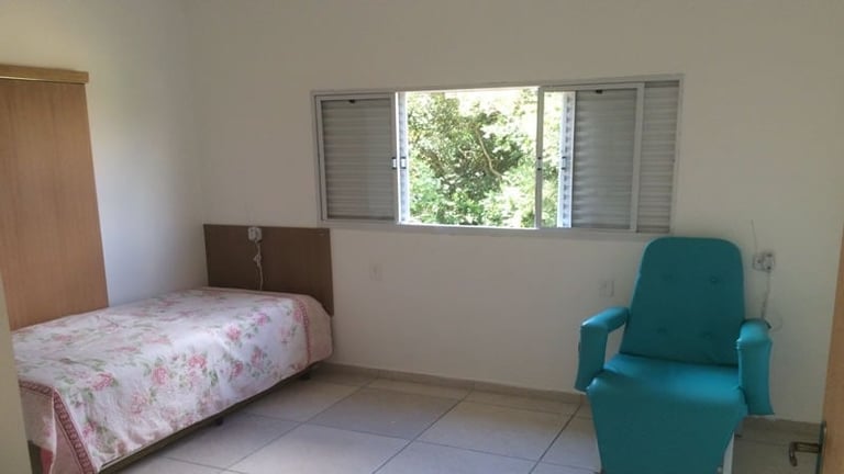A simple bedroom with a single bed with floral bedding, a teal armchair, and a window with blinds overlooking greenery