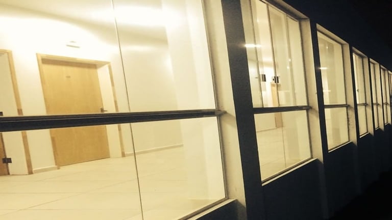 Row of modern glass-walled office cubicles or conference rooms with black frames and cream-colored interior walls, lit from above