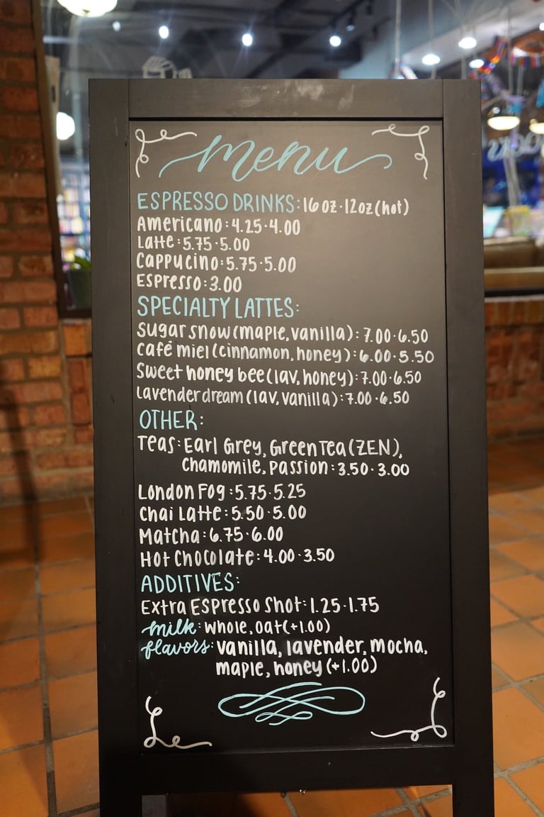 Coffee shop menu board displaying espresso drinks, specialty lattes, teas, and additives with prices written in white chalk