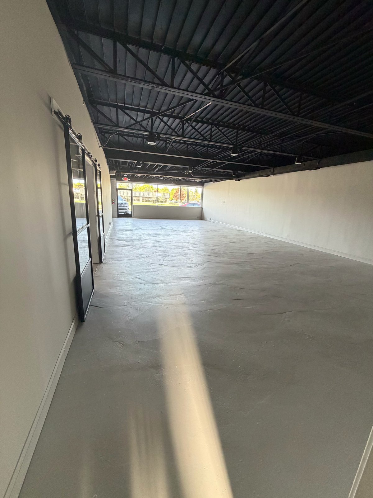 Empty commercial warehouse interior with concrete floor, white walls, metal ceiling structure, and glass storefront windows
