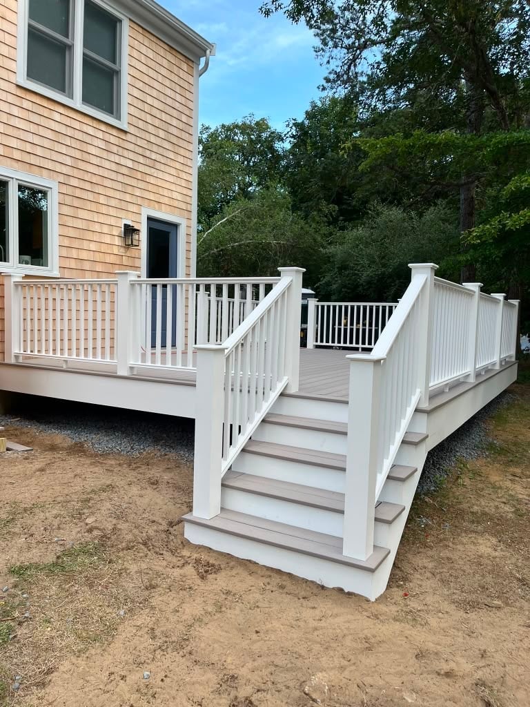 White deck with stairs and railings attached to a beige house with green foliage in background