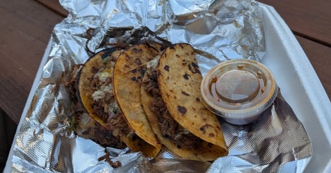 Four birria tacos in a foil-lined container with a small cup of consommé for dipping