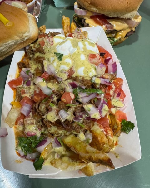 Loaded french fries topped with cheese, meat, onions, tomatoes, and green herbs on a white plate with sandwiches in background
