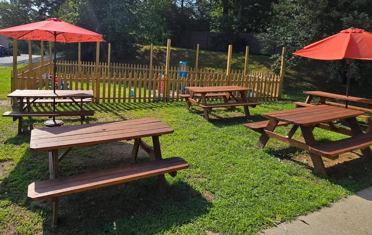 Outdoor park picnic area with wooden tables under red umbrellas, playground equipment and fencing on grass