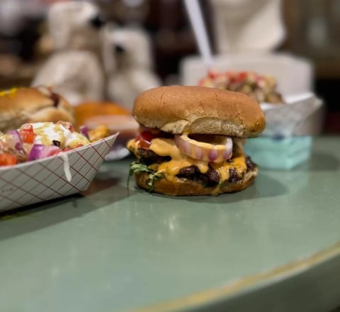 Cheeseburger with melted cheddar and lettuce on a sesame bun, displayed on a light blue counter with side dishes nearby