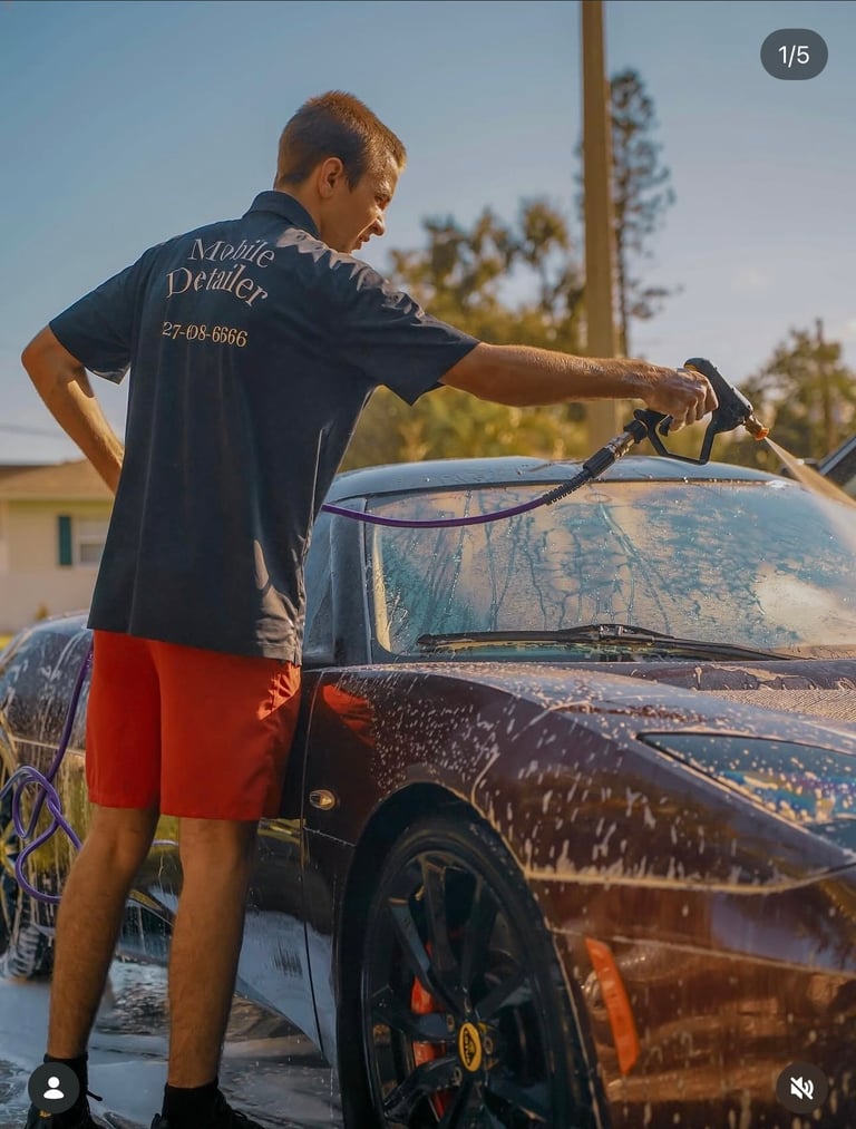 Man washing a dark car with a hose on a sunny day, wearing a navy shirt and red shorts