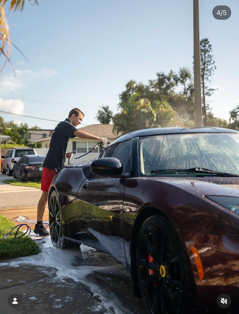 Man washing dark car with hose in driveway on sunny day with residential neighborhood in background