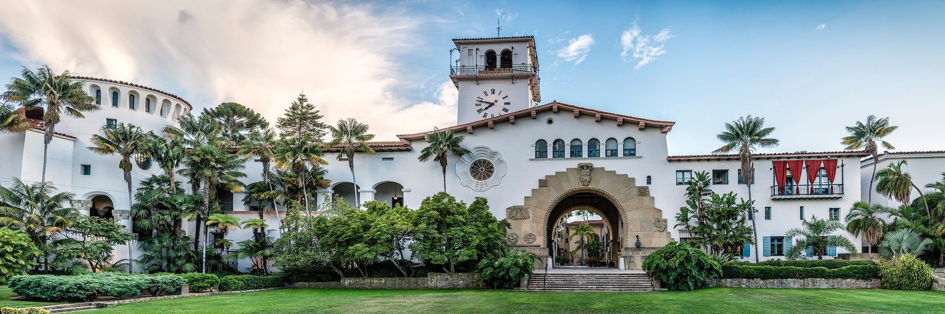 White Spanish Colonial-style building with tower and arched entrance, surrounded by palm trees and green landscaping