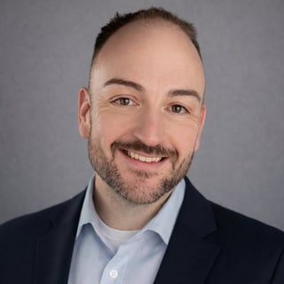 Professional headshot of smiling man with dark beard wearing navy blazer and light blue shirt