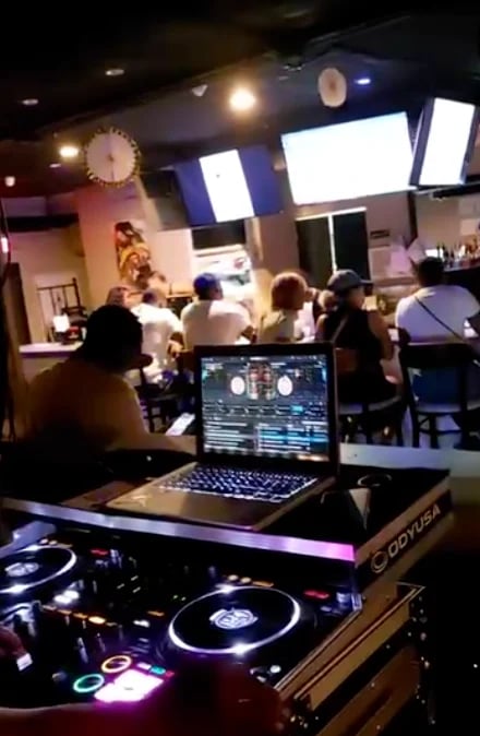 DJ booth with turntables and laptop in foreground, crowd dancing in dimly lit venue with stage lighting