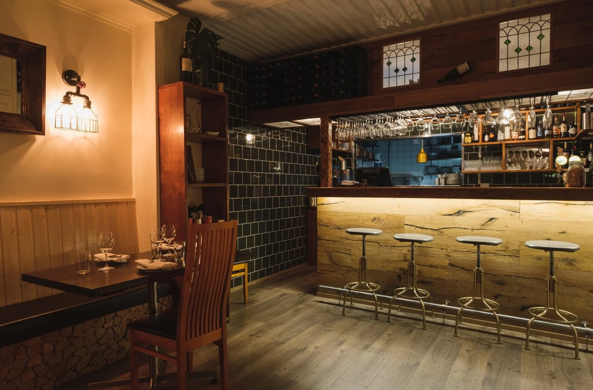 Warm-toned bar interior with booth seating, illuminated counter with bar stools, and shelves of bottles behind the counter