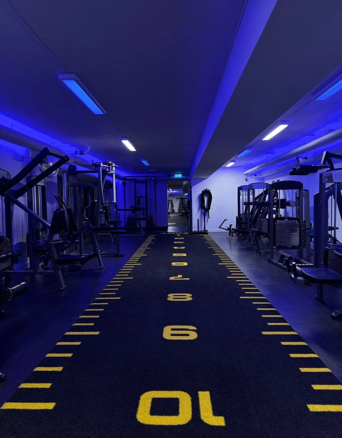 Modern gym interior with blue lighting, numbered yellow floor markings, and exercise equipment lining both sides of the corridor