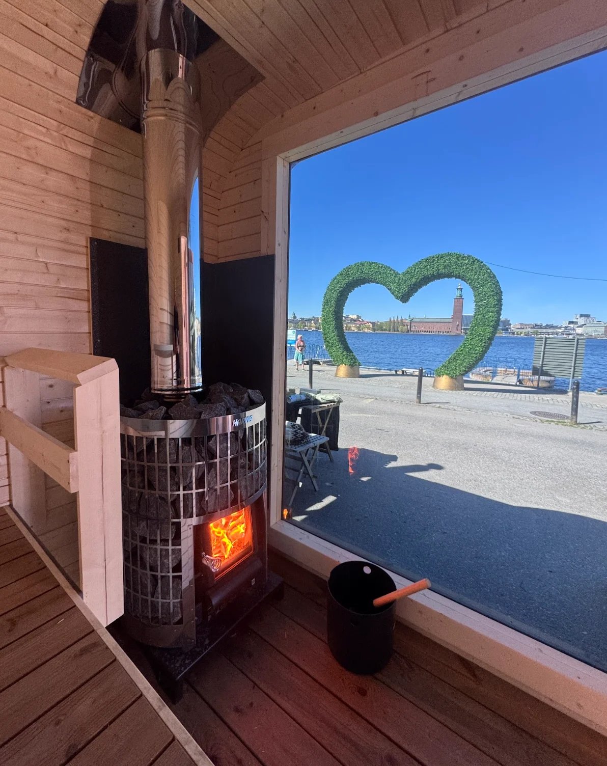 Wood sauna interior with glowing stove, overlooking beach with green heart sculpture and waterfront cityscape