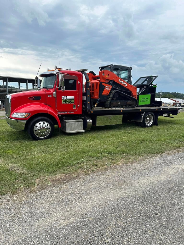 Red Peterbilt truck with orange skid steer loader on flatbed trailer parked in grassy field
