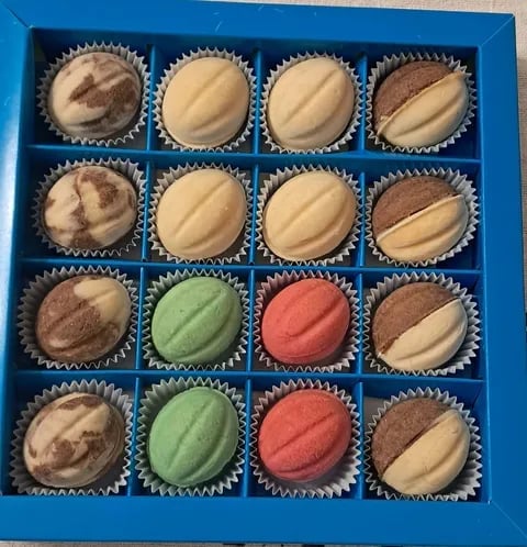 A blue box of 16 assorted macarons in paper cups, featuring vanilla, chocolate, pistachio, and raspberry flavors arranged in rows