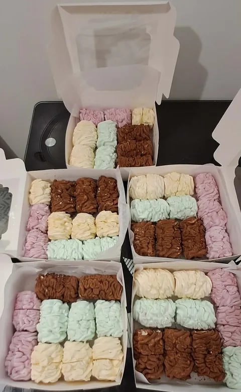 Multiple white boxes containing assorted brownies and rice crispy treats in pink, cream, chocolate, and light blue colors arranged in rows