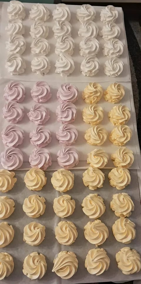 Assorted frosted meringue piped cookies on parchment paper in white, pink, and yellow colors arranged on trays