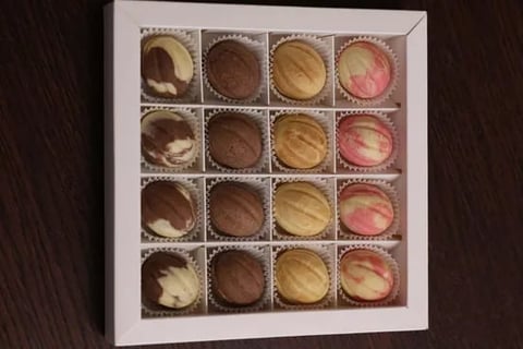 Box of 16 assorted chocolates and bonbons arranged in a 4x4 grid, displaying various flavors and toppings in brown, tan, and pink colors
