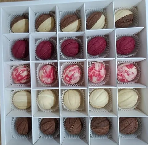 A box of 25 artisan chocolates arranged in a 5x5 grid, featuring various flavors with chocolate, raspberry, and vanilla varieties in white paper liners