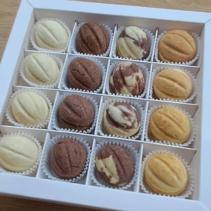 White box containing 16 assorted madeleines including vanilla, chocolate, and swirled varieties in paper cups
