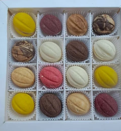 A box of 16 assorted macarons in white paper cups, featuring yellow, pink, chocolate, cream, and tan flavors arranged in a 4x4 grid