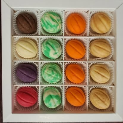 A box of 16 colorful macarons arranged in a 4x4 grid, featuring flavors in brown, green, orange, yellow, purple, and red varieties in paper liners