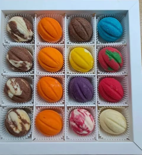 Box of 16 colorful macarons in white compartments, including chocolate, orange, blue, yellow, pink, and purple flavors