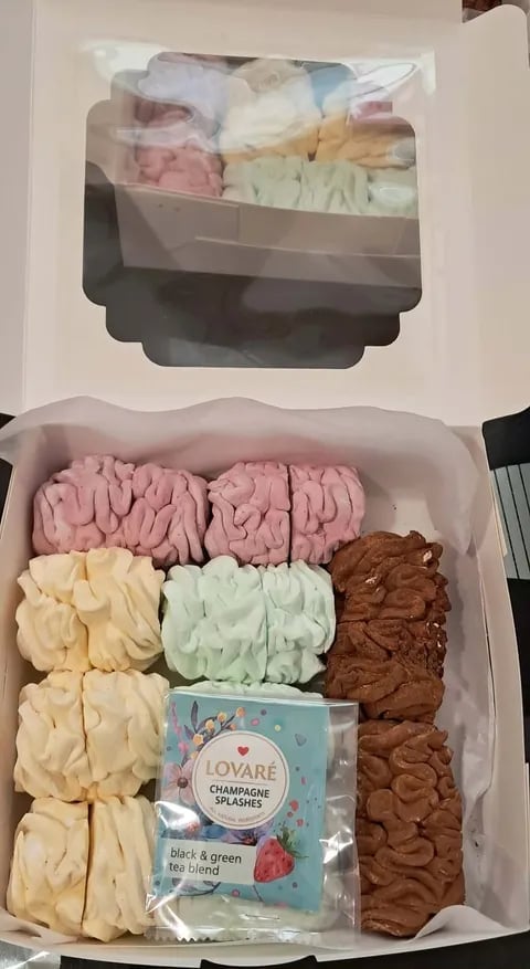 Open box containing assorted colorful meringue cookies in pink, cream, green, and brown, with a small tea blend packet in the center