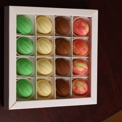 Box of 16 colorful macarons arranged in a 4x4 grid with green, yellow, brown, and pink varieties