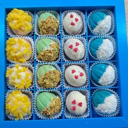 A blue box containing 16 assorted decorated truffles in four rows, featuring yellow, pistachio, white with red dots, and turquoise designs