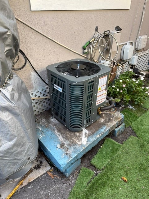 Outdoor HVAC air conditioning unit mounted on a concrete pad next to a house with surrounding pipes and cables
