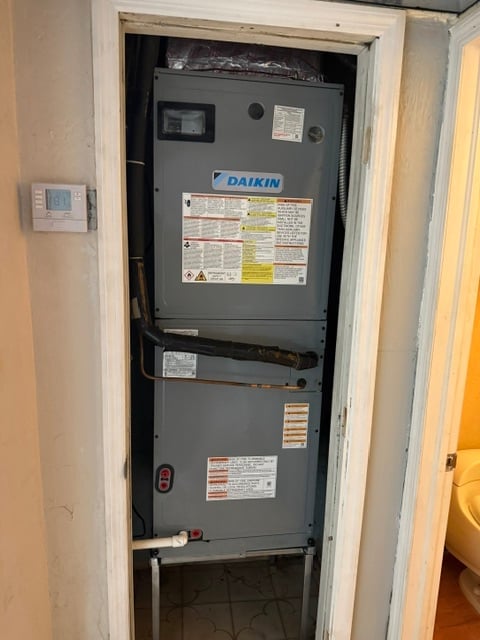Daikin indoor air conditioning unit mounted in a utility closet with instruction labels and thermostat visible on wall.