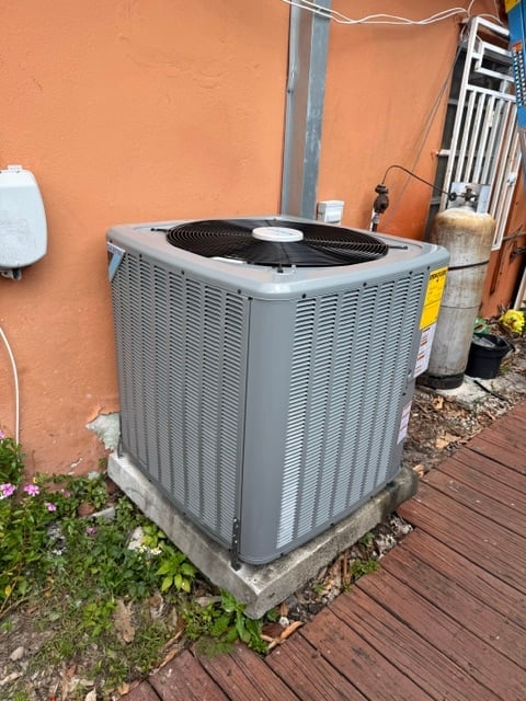 Gray HVAC air conditioning unit installed against an orange wall with concrete foundation, yellow caution label visible