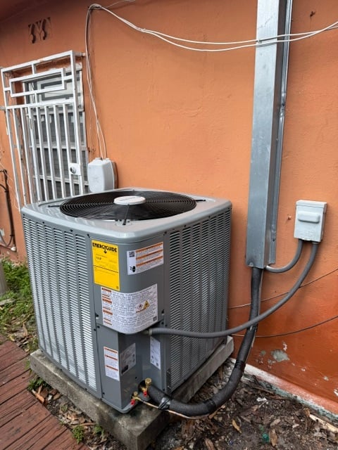 Outdoor air conditioning unit installed against an orange exterior wall with power lines and electrical connections visible.