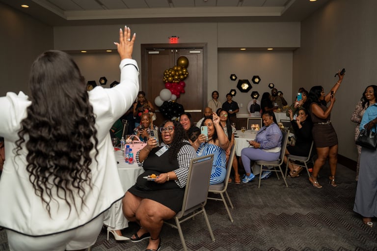 Woman with long dark hair waving from behind, addressing a crowd at an indoor holiday celebration with decorations