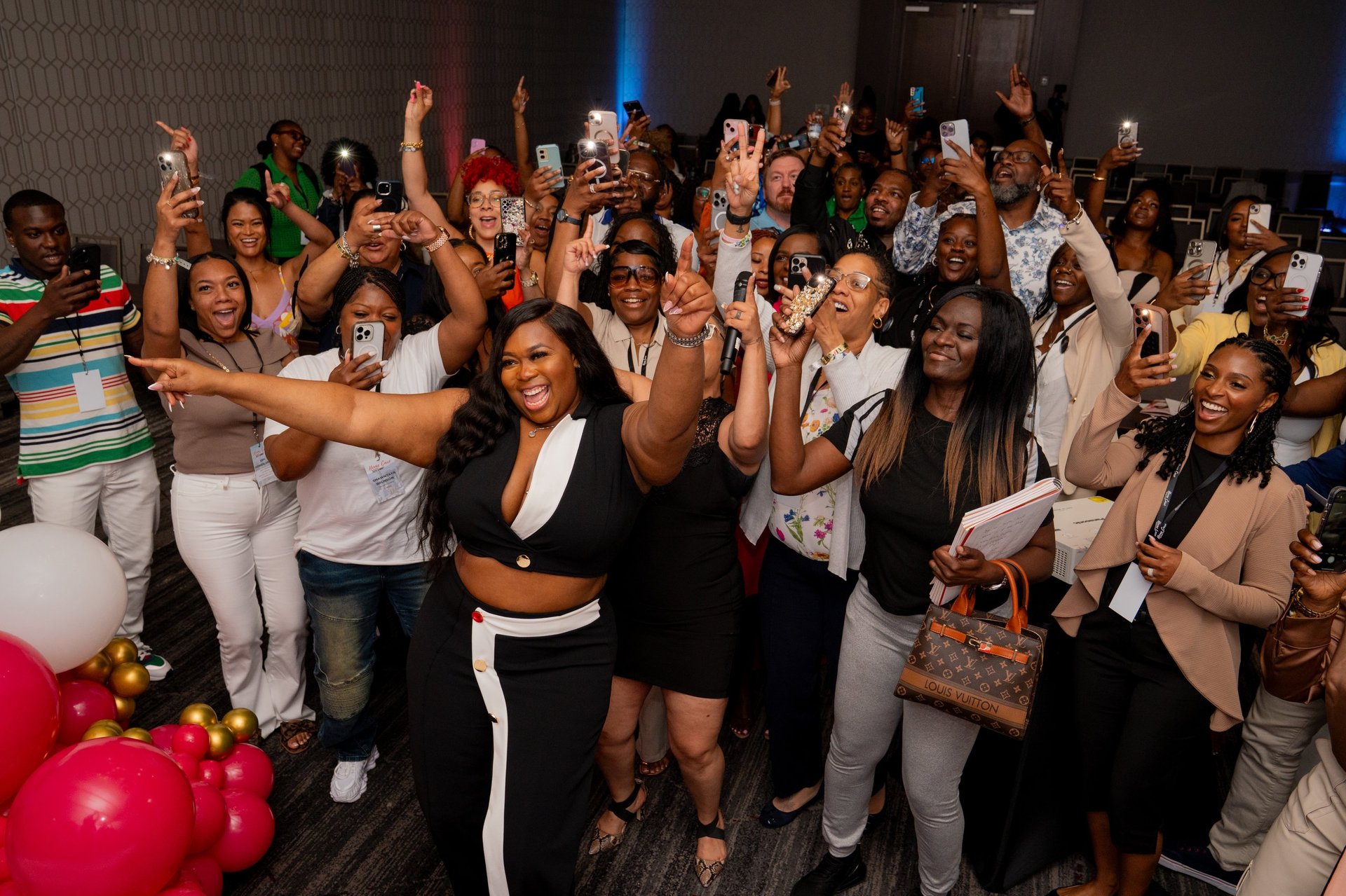 Large group of people celebrating at an indoor event with raised arms, drinks, and festive decorations including pink balloons.