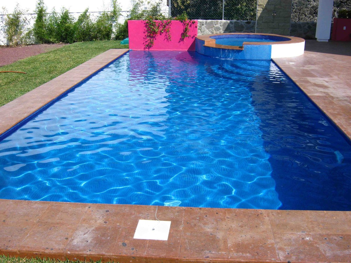 Alberca rectangular con agua azul brillante, elementos rosas decorativos, jacuzzi blanco y deck de ladrillo en jardín