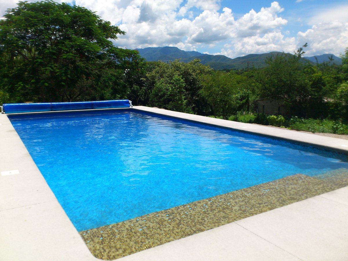 Alberca rectangular moderna con agua azul brillante, deck de concreto blanco y montañas con bosque al fondo en día soleado