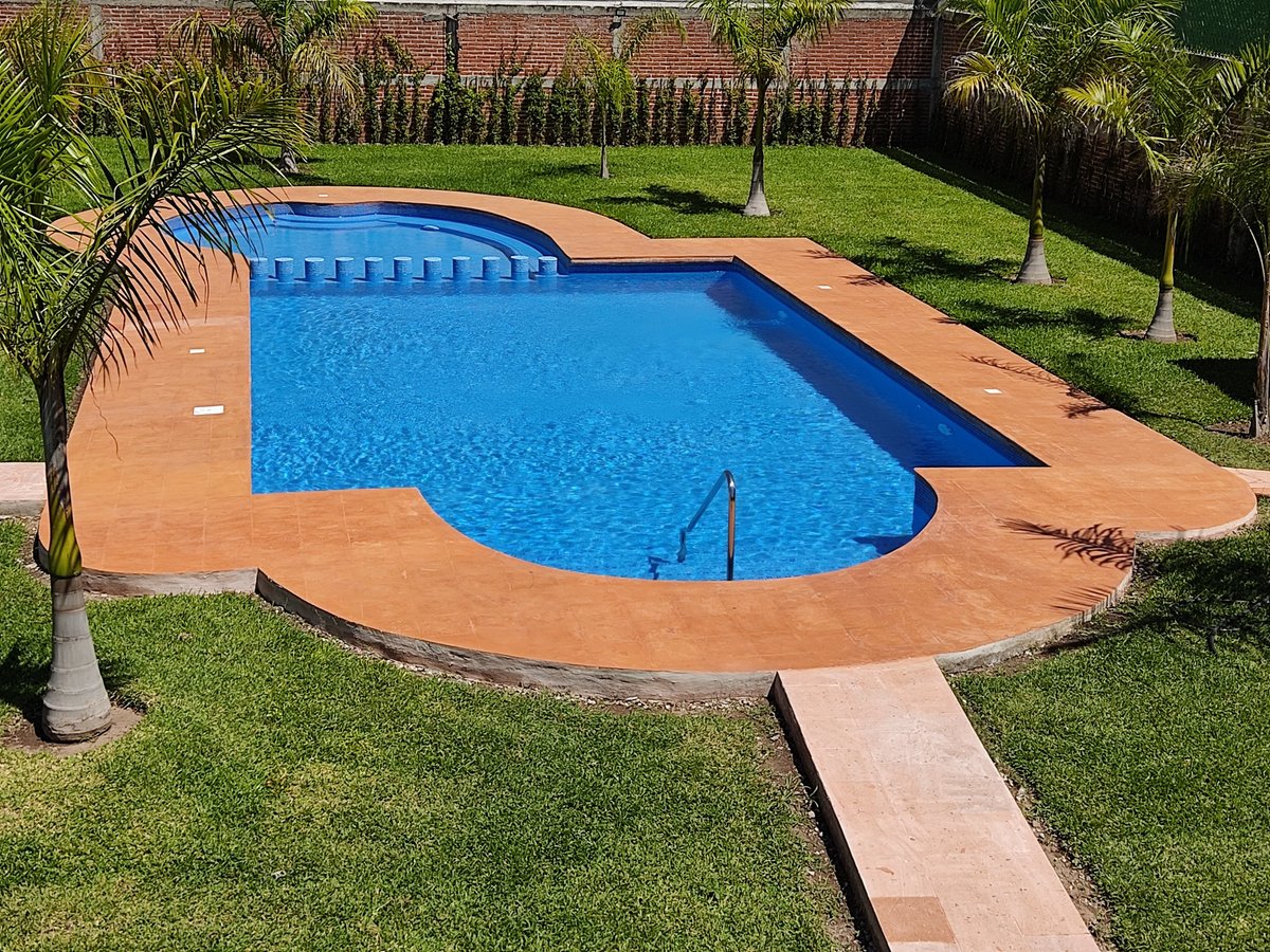 Vista aérea de alberca curva con agua azul, deck de terracota, rodeada de pasto verde cuidado y palmeras