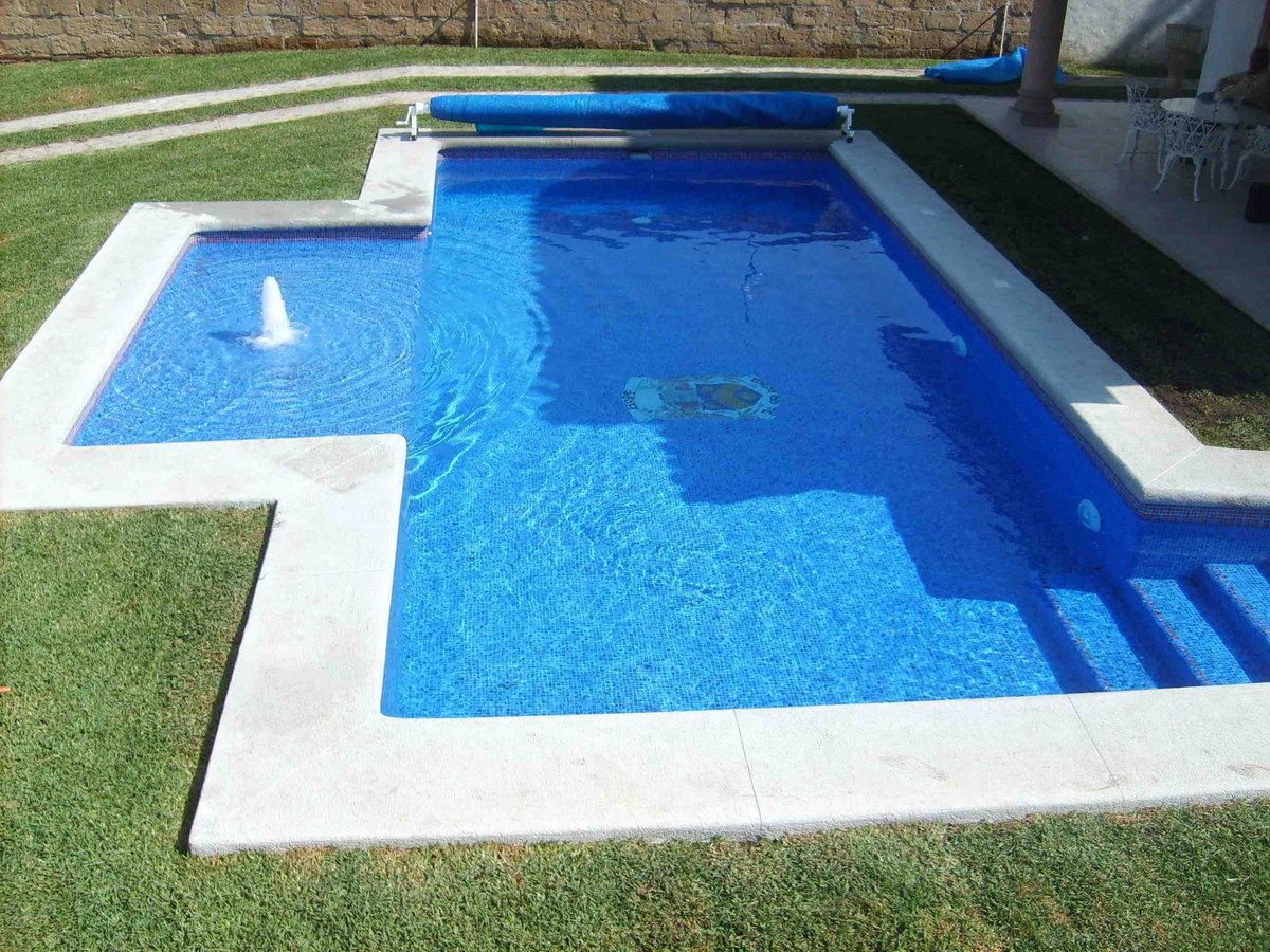 Alberca rectangular en jardín con agua azul, deck de concreto blanco y enrollador de cubierta azul rodeada de pasto