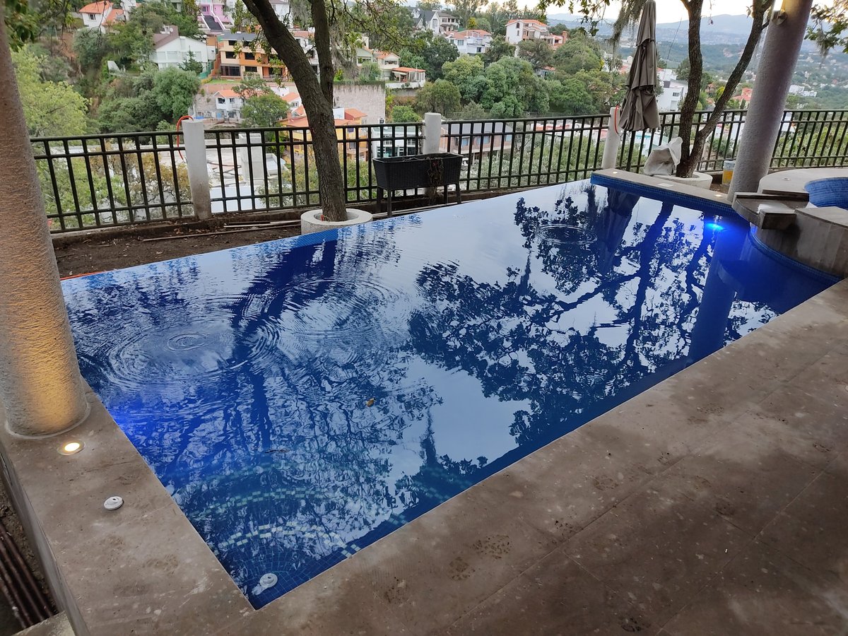 Alberca rectangular con agua azul reflejando árboles y cielo, rodeada de barandales metálicos y casas en ladera