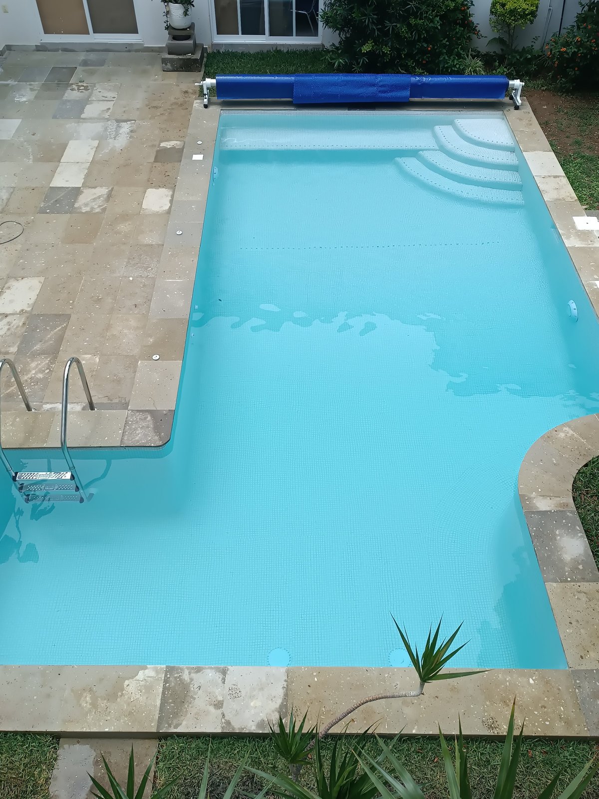 Vista aérea de alberca rectangular con agua turquesa brillante y enrollador de cubierta azul, rodeada de patio de piedra