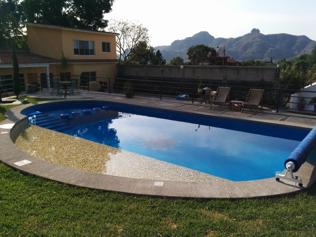 Alberca rectangular grande con agua azul brillante en jardín con casa amarilla, tumbonas y montañas al fondo