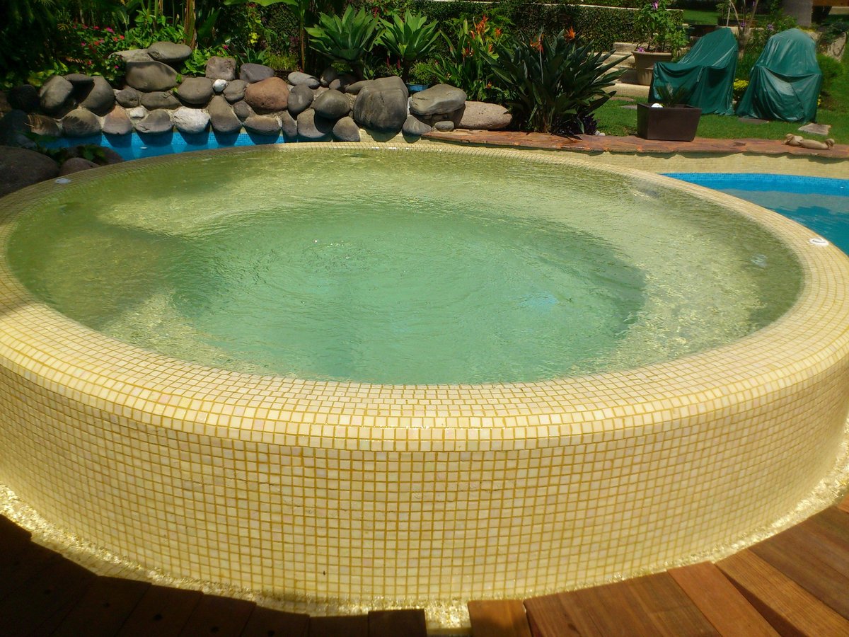 Alberca redonda en jardín con borde de azulejo amarillo y agua turquesa, rodeada de rocas y paisajismo
