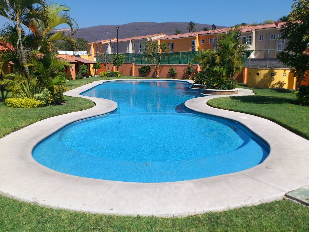 Dos albercas curvas con agua azul rodeadas de deck blanco, pasto verde, palmeras y casas residenciales coloridas al fondo