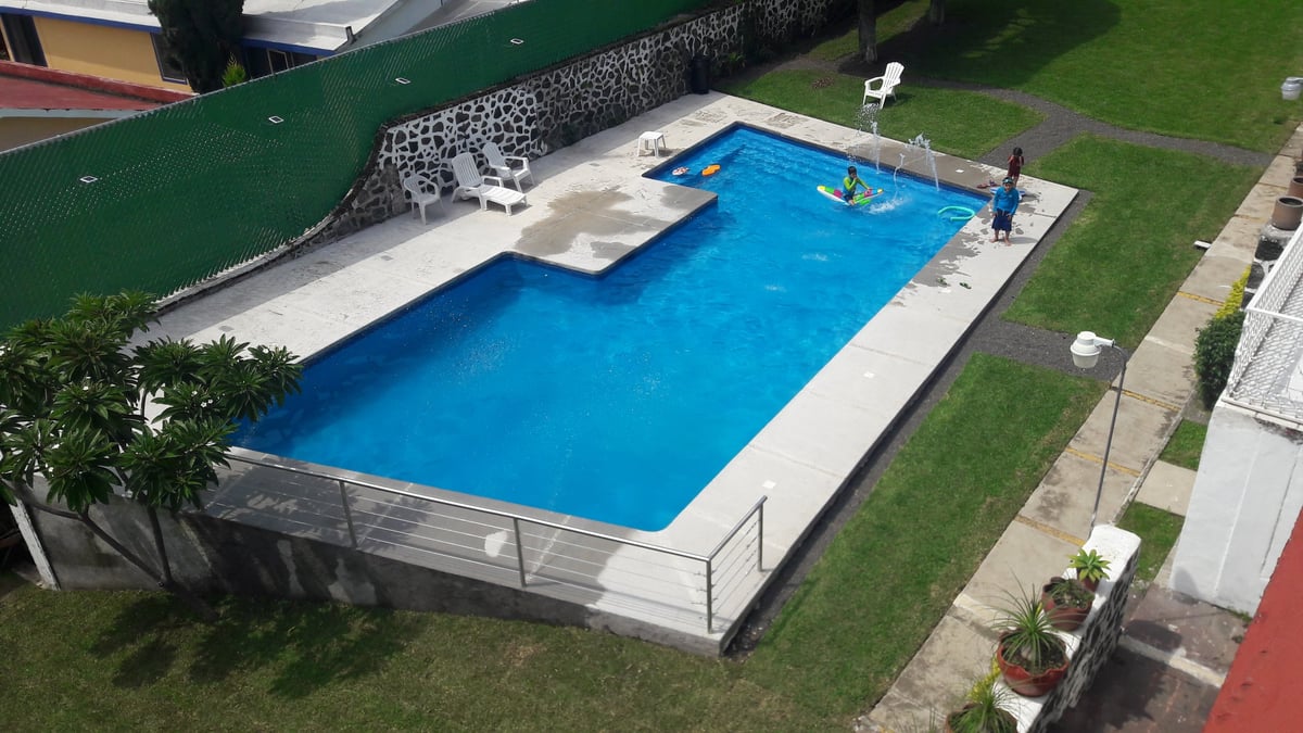 Vista aérea de alberca rectangular con agua azul brillante, deck blanco, jacuzzi y jardín verde en casa residencial