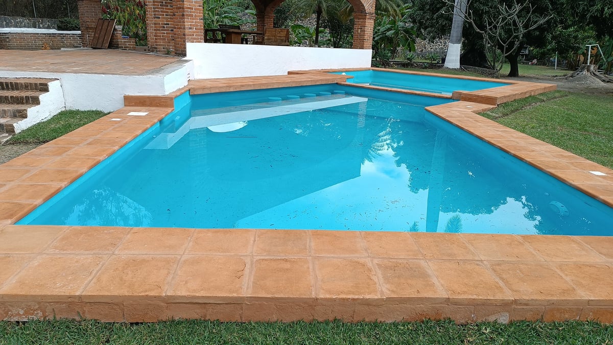 Alberca rectangular con agua azul brillante, deck de loseta terracota y casa blanca de alberca, rodeada de árboles y jardín con paisajismo